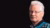 A portrait of Professor Peter van Bergeijk. In the background is a black backdrop. He wears a thin-framed pair of glasses and a pattern blue jacket.