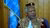 Mohamed Salih in red and gold patterned robe and kufi, before wooden wall and blue flag.