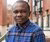Photo of a young Black man in a blue plaid shirt standing outside brick townhouses