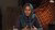 Woman in teal hijab and embroidered top speaking, patterned textiles in background.