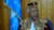 Mohamed Salih in red and gold patterned robe and kufi, before wooden wall and blue flag.