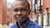 Photo of a young Black man in a blue plaid shirt standing outside brick townhouses