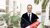 Smiling man in graduation gown holding oversized red diploma tube in atrium