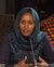 Woman in teal hijab and embroidered top speaking, patterned textiles in background.