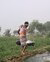 A farmer in Bangladesh carrying water to irrigate crops in a lush green field under a clear sky.