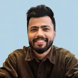 In this portrait, Abhimanyu smiles for the camera. The background of the photo is a light blue, textured shade. Abhimanyu wears a dark brown button up shirt.