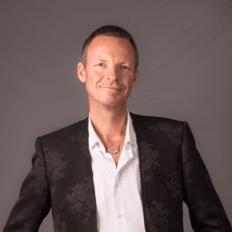In this portrait photo, Niels van Doesum smirks at the camera. He wears a black jacket on top of a white button up shirt, complete with a unique silver necklace.