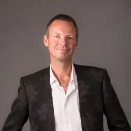 In this portrait photo, Niels van Doesum smirks at the camera. He wears a black jacket on top of a white button up shirt, complete with a unique silver necklace.
