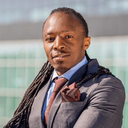 Photograph of man with locks in gray suit and brown tie standing before glass building