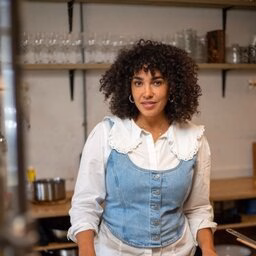 Women wearing a blue top over white top with curly hair standing in kitchen